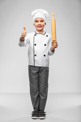 cooking, culinary and profession concept - happy smiling little boy in chef's toque and jacket with rolling pin showing thumbs up over grey background