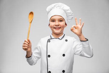 cooking, culinary and profession concept - happy smiling little boy in chef's toque and jacket with wooden spoon showing ok gesture over grey background