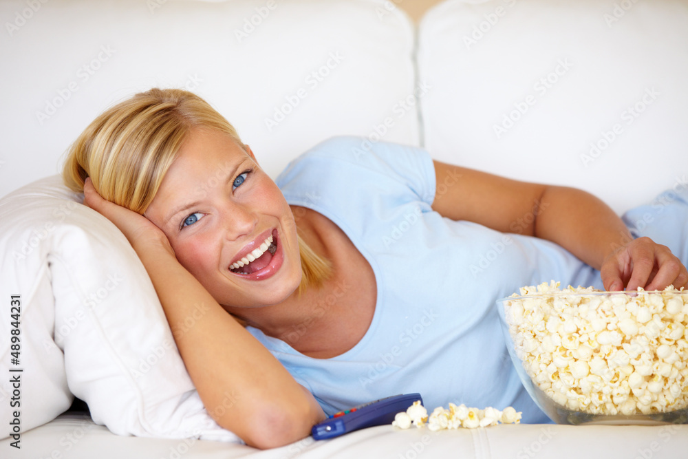 Poster Hey, its my favorite show. Young woman lying on the couch and watching TV while eating popcorn.