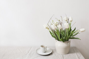Feminine Easter, spring still life scene. Cup of coffee and floral bouquet. White ceramic vase on table. White tulips flowers, cherry tree branches on linen tablecloth. Styled interior. Copy space.