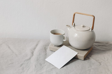 Breakfast still life scene. Cup of coffee, ceramics tea pot on old book. Blank greeting card, invitation mockup. Beige linen tablecloth. White wall backgroound. Neutral morning lifestyle composition.