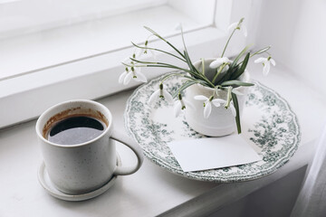 Winter, spring still life scene. Cup of coffee on windowsill. Vase of snowdrops flowers on vintager green decorative plate. Feminine floral composition with galanthus plants. Blurred background.