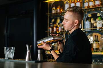 Confident barman in black clothes mixing ingredients of alcoholic cocktail by shaking shaker standing behind bar counter, on blurred background of shelves with alcoholic drinks, selective focus.