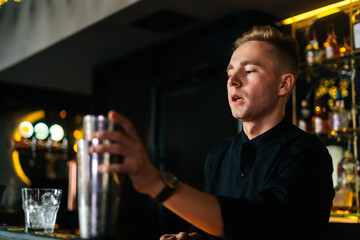 Close-up of focused bartender preparing refreshing alcoholic cocktail standing behind bar counter in modern dark nightclub, on blurred background of shelves with different alcoholic drinks.
