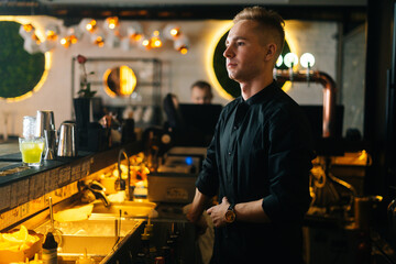 Medium shot portrait of handsome young barman standing near bar counter waiting for customers orders on blurred background of modern nightclub with dark interior, selective focus.