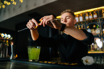 Close-up low-angle view of young bartender male squeezing out piece of orange peel with straw and decorating cocktail, standing behind bar counter in modern nightclub with dark interior.