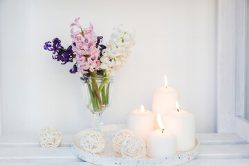 Obraz na płótnie Canvas A bouquet of white, pink, blue cut hyacinth a in a small white corrugated vase and three large burning candles on a round tray are on a beige table. Place for text