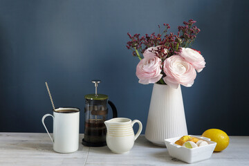 Coffee cup and bouquet of pink Persian buttercups on wooden table. mock up background for design. Yellow and white macaroons in a white porcelain bowl. Dark blue background. Copy space