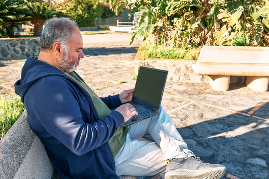Handsome bearded mature man with laptop computer working outdoors while sitting on bench at the seaside. Modern lifestyle, connection, business, freelance work concept.
