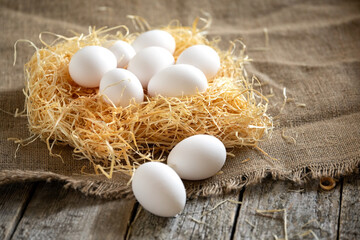 White chicken eggs in straw or hay nest on a burlap sackcloth on rough wooden country table.Still...
