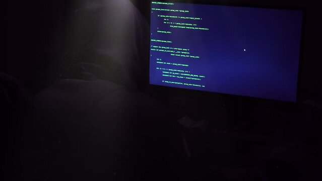 Computer Hacker In Hooded Shirt Using Computer Keyboard And Coding Ransomware Attack To Steal Personal Data In Cyber Attack. Hacker's Hands Typing Malicious Code Into Keyboard On Large Screen.