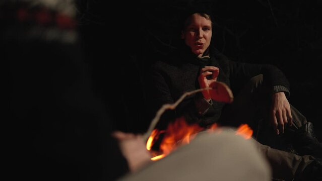 Two friends are chatting around a campfire in the forest. Two brothers have fun and talk about the good life. Camping in nature in the forest in the warm season. Positive men spend time outdoors