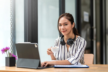 Charming young Asian businesswoman sitting smiling working drinking coffee and using laptop on the desk in the office.