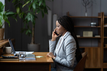 African businesswoman provide help consulting client distantly by mobile phone call sit at desk with laptop computer in office. Busy successful employee has conversation with customer on smartphone