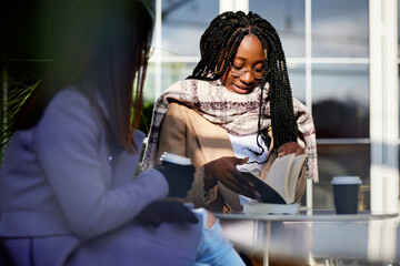 Two multicultural girls sitting outside of cafe, drinking coffee and reading books. Students learning.