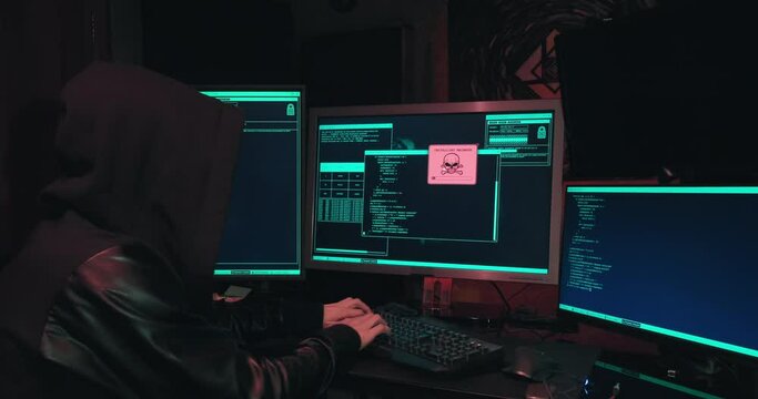 Hacker in a hood sits in front of computer screens and hacks databases. The concept of cyberterrorism, server hacking and cyberattacks. The room is dark, a person is typing code into the command line