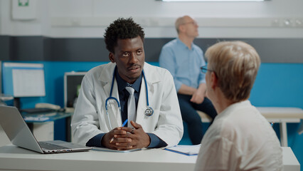 Adult specialist consulting senior woman sitting at desk writing checkup notes on files in medical office. Old patient receiving professional advice for man on bed in background