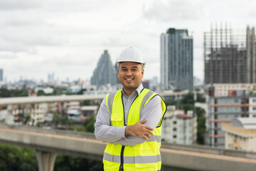 Asian engineer man or architect looking forward with white safety helmet in city construction site . Standing on rooftop building construction at capital.