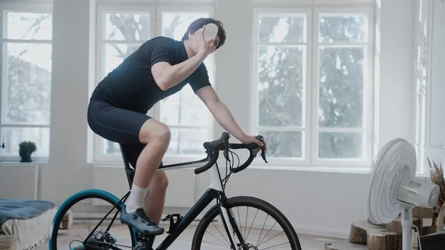 Side view of a cyclist training on an exercise bike and drinking water at home. Man cycling on smart trainer wearing cycling shoes, staying at home during pandemic. High quality 4k footage