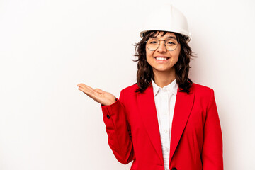 Young architect hispanic woman with helmet isolated on white background showing a copy space on a palm and holding another hand on waist.