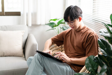 Fototapeta premium Technology Concept The male with his casual T-shirt and jeans sitting comfortably on the wooden chair and doing touchscreen for checking the web browser on the iPad