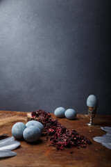 DIY blue painted easter eggs, one egg in egg cup, dry Hibiscus tea and silver decorative feathers on wooden background, copy space, vertical shot