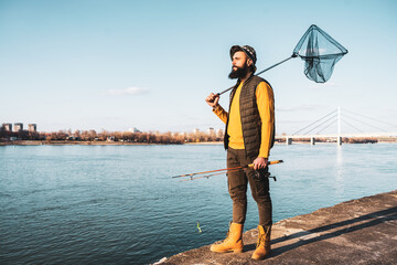 Image of fisherman holding fishing rod  and fishing net while standing by a river.