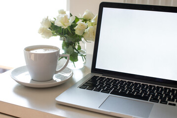 Laptop with white blank screen on white work table. White interior home office concept. Female office desk workspace with white roses and cup of coffee. Close up on screen laptop. Mock up laptop