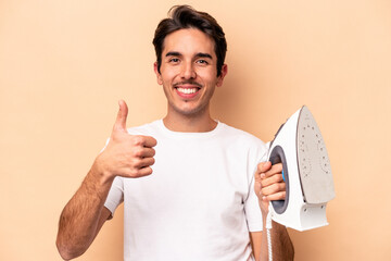 Young caucasian man holding an iron isolated on beige background smiling and raising thumb up
