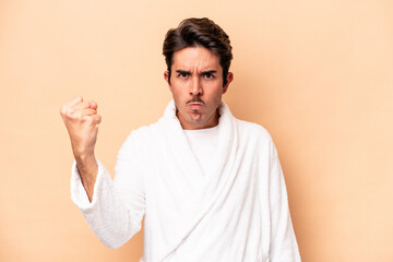 Young caucasian man wearing a bathrobe isolated on beige background showing fist to camera, aggressive facial expression.