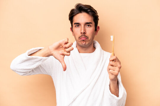 Young caucasian man wearing a bathrobe holding a toothbrush isolated on beige background showing a dislike gesture, thumbs down. Disagreement concept. - Powered by Adobe