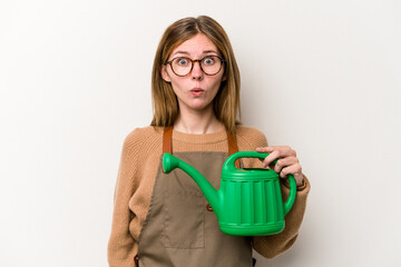 Young gardener woman holding a sprinkler isolated on white background shrugs shoulders and open eyes confused.