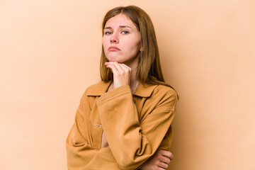 Young English woman isolated on beige background suspicious, uncertain, examining you.
