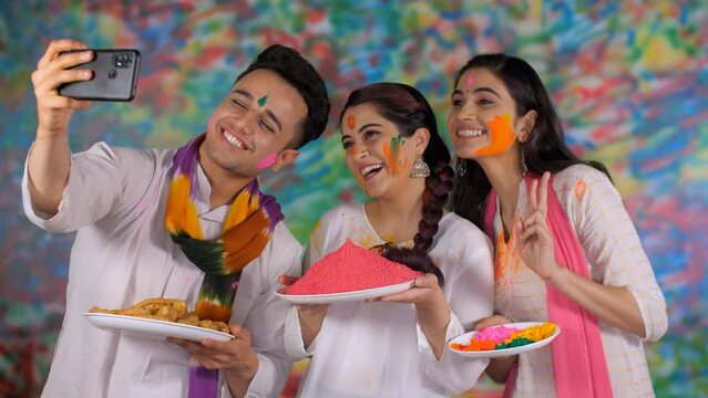College friends wearing a white Kurta and a scarf enjoy clicking a selfie on Holi - festive season  leisure time  togetherness and bonding. Cheerful siblings standing with a plate of Gulal and trad...