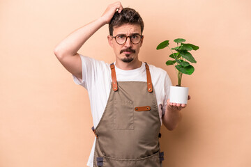 Young caucasian gardener man holding a plant isolated on beige background being shocked, she has...