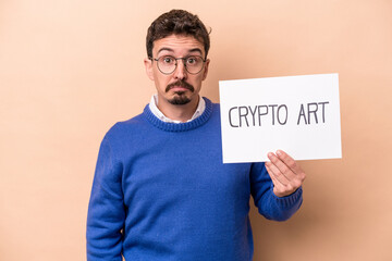 Young caucasian man holding a crypto art placard isolated on beige background shrugs shoulders and...
