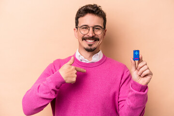 Young caucasian man holding batteries isolated on beige background person pointing by hand to a shirt copy space, proud and confident