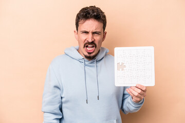 Young caucasian man holding a puzzle isolated on beige background screaming very angry and aggressive.