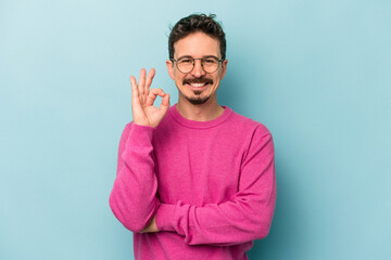 Young caucasian man isolated on blue background winks an eye and holds an okay gesture with hand.