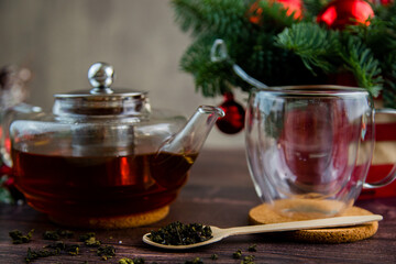 A transparent teapot with tea stands on the table, a transparent mug stands on a wooden stand next to it, a wooden spoon lies next to it and tea is scattered on a dark background. High quality photo