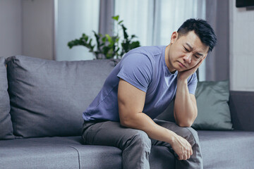 Upset Asian man at home, tired and depressed sitting on the couch in the living room
