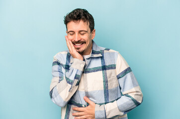 Young caucasian man isolated on blue background laughs happily and has fun keeping hands on stomach.