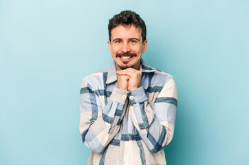 Fototapeta premium Young caucasian man isolated on blue background keeps hands under chin, is looking happily aside.