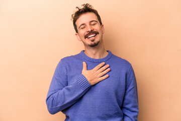Young caucasian man isolated on beige background laughs out loudly keeping hand on chest.