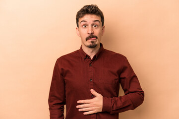 Young caucasian man isolated on beige background touches tummy, smiles gently, eating and satisfaction concept.