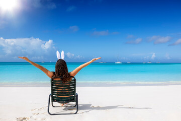 Easter travel concept with a happy woman with bunny ears sitting on a tropical beach with...