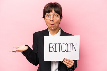 Young hispanic business woman holding a bitcoin placard isolated on pink background shrugs...