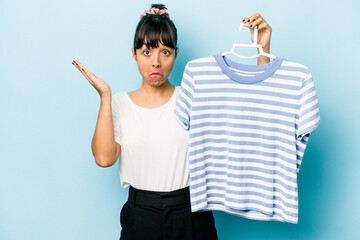 Young hispanic woman holding a t-shirt on a hanger isolated on blue background shrugs shoulders and...
