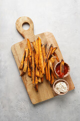 Healthy homemade oven baked sweet potato fries on a baking parchment, wooden board, with ketchup sauce and mayonnaise