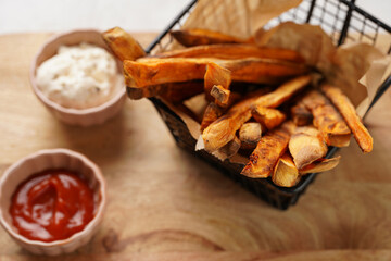 Healthy homemade oven baked sweet potato fries in a black deep frying basket net grid, wooden board, with ketchup sauce and mayonnaise